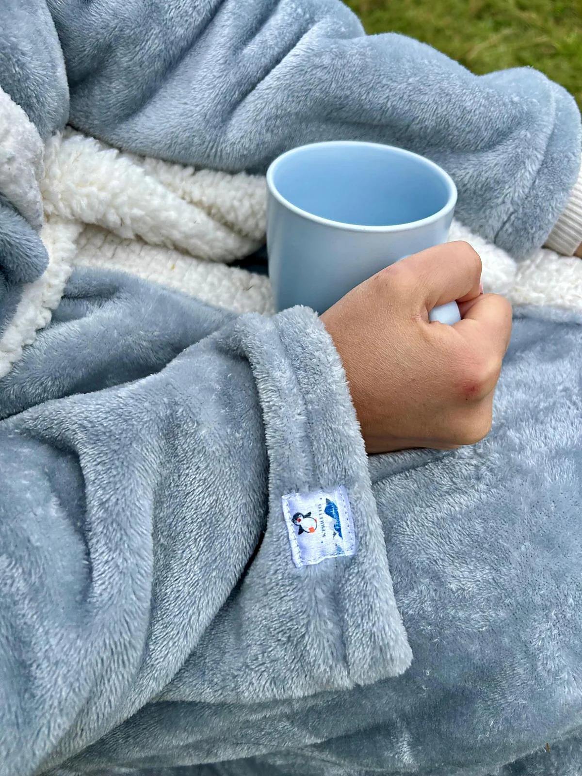 Mano sosteniendo una taza sobre una prenda K'priletto gris de felpa con forro de borrego y etiqueta del pingüino